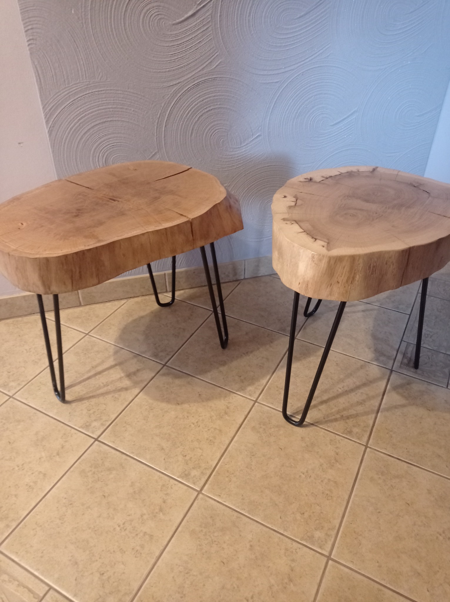 Dwa małe stoliki kawowe wykonane z plastrów drewna o nieregularnym kształcie, osadzone na czarnych, metalowych nóżkach typu hairpin, ustawione na podłodze z płytek ceramicznych w jasnym odcieniu...
