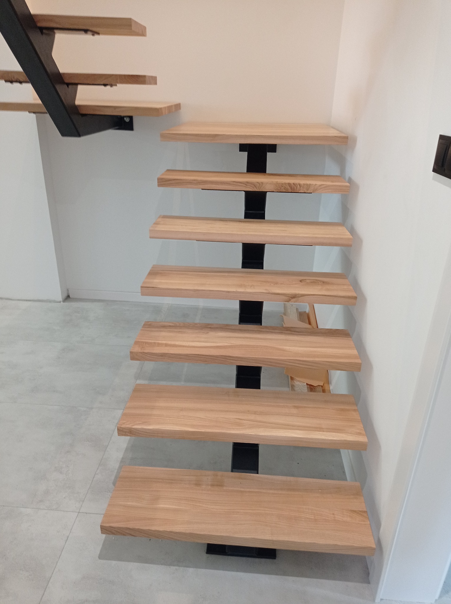 Nowoczesne schody wspornikowe z drewnianymi stopniami i czarną metalową konstrukcją nośną, widoczne z boku na tle białej ściany i szarej podłogi.