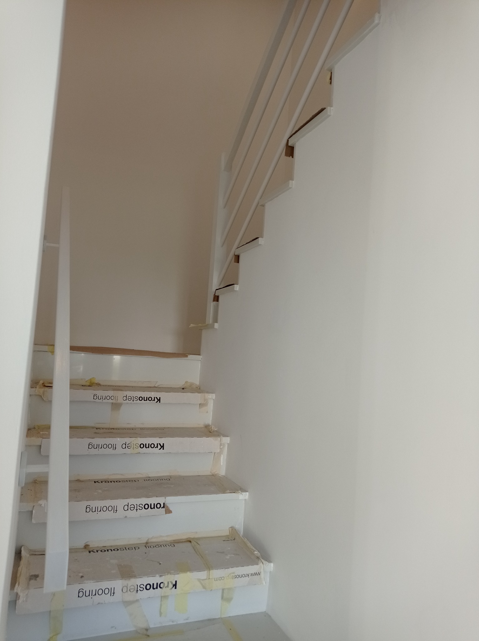 Białe schody z barierką w trakcie montażu, zabezpieczone kartonem i taśmą, widoczne logo Kronostep flooring na zabezpieczeniach.