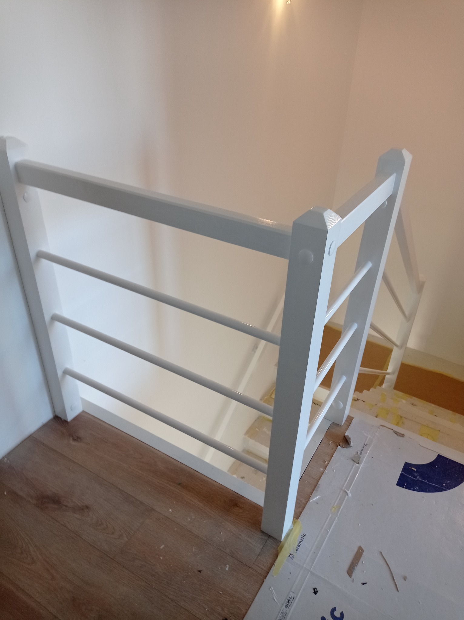 Biała balustrada schodowa z drewnianymi szczeblami, widok z góry na drewnianą podłogę i zabezpieczone schody w trakcie montażu.