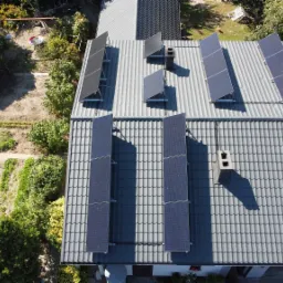 Widok z drona na dach domu z szarej dachówki, na którym zamontowano panele fotowoltaiczne w układzie pionowym i poziomym, obok komin z cegły oraz bujna zieleń ogrodu.