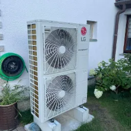 Biała zewnętrzna jednostka pompy ciepła LG Therma V, umieszczona na betonowym fundamencie przy białej ścianie domu, z zielonym wężem ogrodowym zwiniętym na felgę w tle i kwitnącymi hortensjami obok.