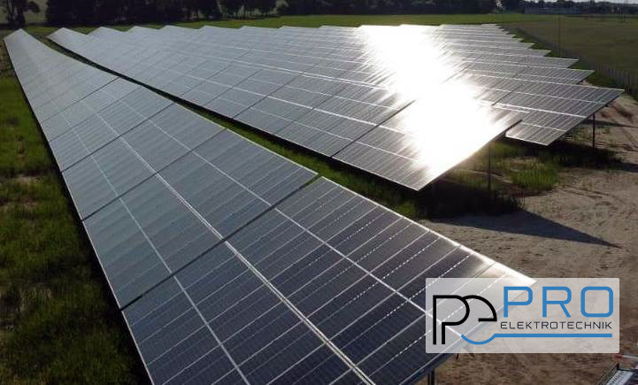 Rozległa farma paneli słonecznych na trawiastym terenie, odbijająca intensywne promienie słoneczne, z logo firmy w prawym dolnym rogu.