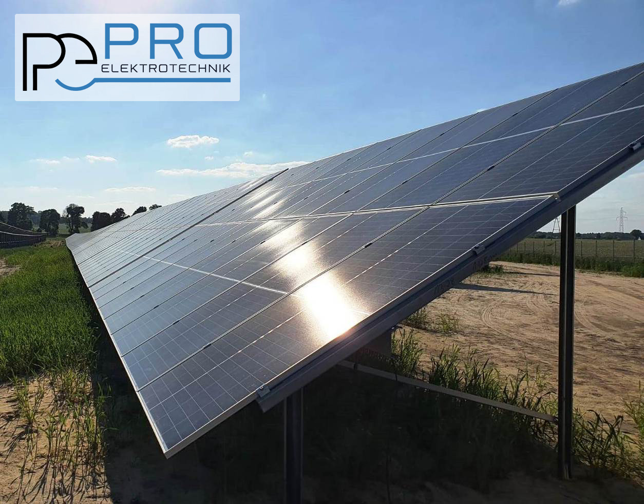 Rozległa instalacja paneli słonecznych z logo firmy w rogu, odbijająca promienie słoneczne na tle błękitnego nieba i polnego krajobrazu.