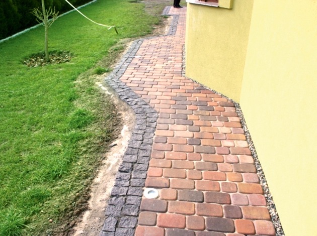 Ścieżka z kostki brukowej w odcieniach czerwieni i brązu, obrzeże z ciemnej kostki granitowej, widok z perspektywy pieszego, trawnik po lewej stronie, ściana budynku w kolorze żółtym po prawej...