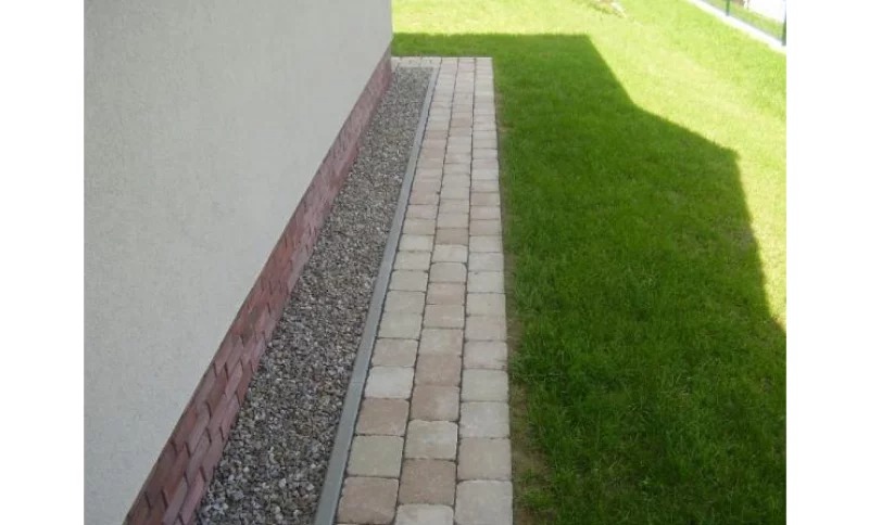 Jasna kostka brukowa ułożona wzdłuż ściany domu z obrzeżem i pasem żwiru, granicząca z trawnikiem w słoneczny dzień.