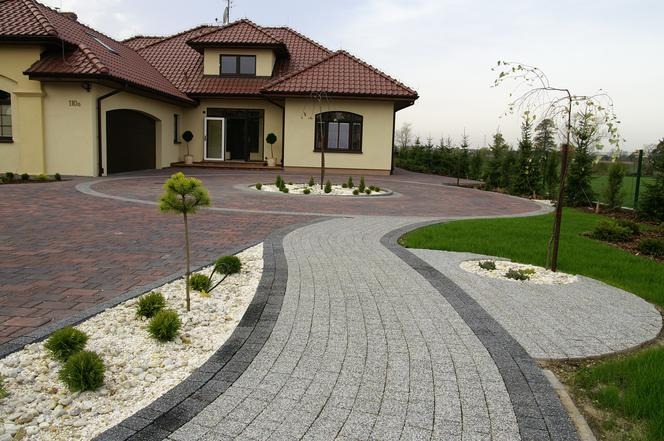 Szeroki, kręty podjazd z kostki brukowej w odcieniach szarości i czerwieni prowadzący do domu z beżową elewacją i czerwoną dachówką, z elementami dekoracyjnymi z białych kamieni i niskiej zieleni.