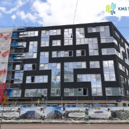 Budynek apartamentowy Starter we Wrocławiu w trakcie budowy, widoczne rusztowania częściowo zasłonięte banerami reklamowymi, pracownik w żółtym kasku na rusztowaniu, elewacja z ciemnej cegły...