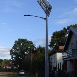 GWARANT Oświetlenia - Hybrydowa latarnia uliczna z panelem solarnym i małą turbiną wiatrową o czerwonych łopatkach, zainstalowana przy drodze w obszarze zabudowanym.