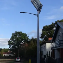 Hybrydowa latarnia uliczna z panelem solarnym i małą turbiną wiatrową o czerwonych łopatkach, zainstalowana przy drodze w obszarze zabudowanym.