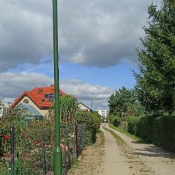 GWARANT Oświetlenia - Nowoczesna, zielona lampa solarna LED oświetlająca ścieżkę w ogrodzie działkowym, widoczna z bliska na tle błękitnego nieba z chmurami i domków z czerwonymi dachami.