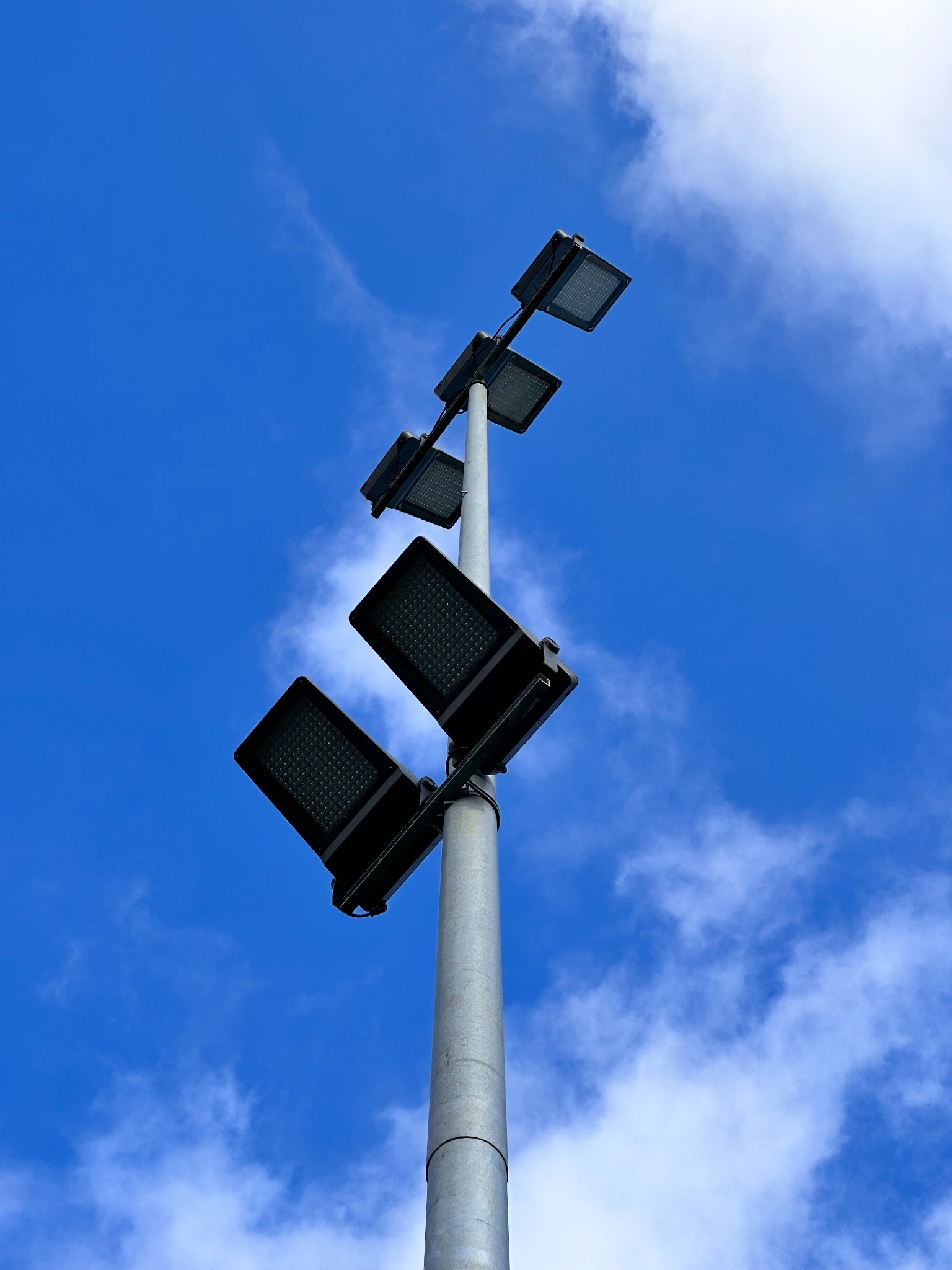 Słup oświetleniowy z czterema czarnymi reflektorami LED, skierowany ku niebu z chmurami.