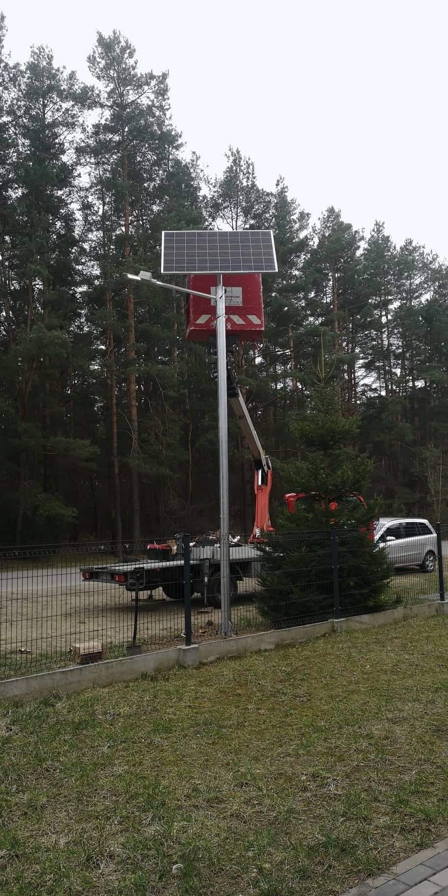 Montaż lampy ulicznej z panelem solarnym na tle lasu, widoczny podnośnik koszowy i samochód dostawczy.