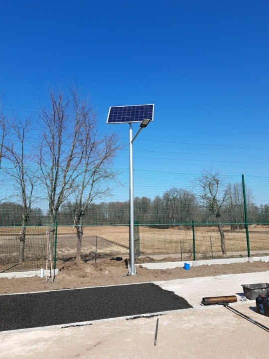 Słup oświetleniowy z panelem solarnym na terenie sportowym, widoczne narzędzia i materiały do wykończenia nawierzchni.