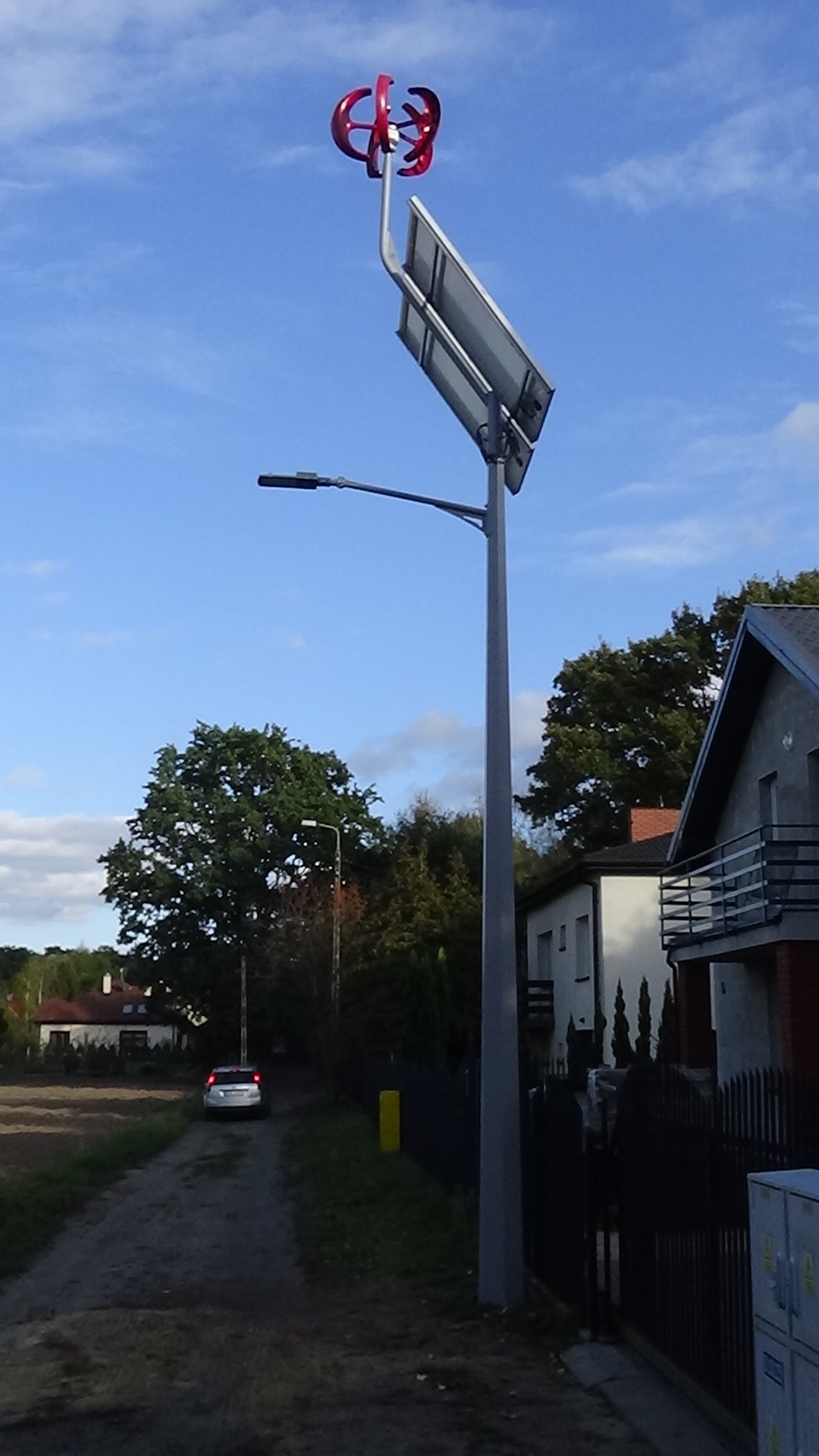 Hybrydowa latarnia uliczna z panelem solarnym i małą turbiną wiatrową o czerwonych łopatkach, zainstalowana przy drodze w obszarze zabudowanym.
