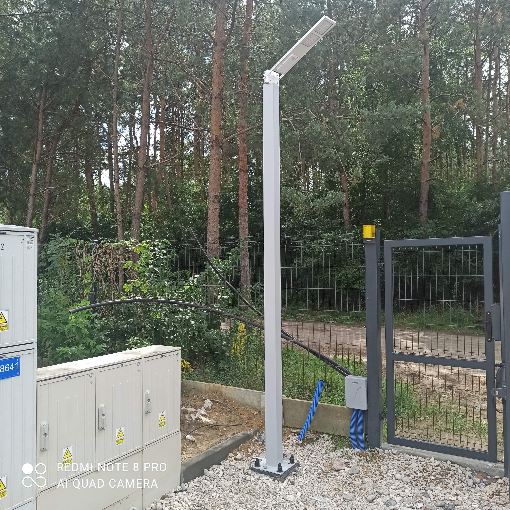 Nowoczesna, biała lampa LED uliczna, zamontowana na posesji z metalową bramą i skrzynkami elektrycznymi w tle lasu.