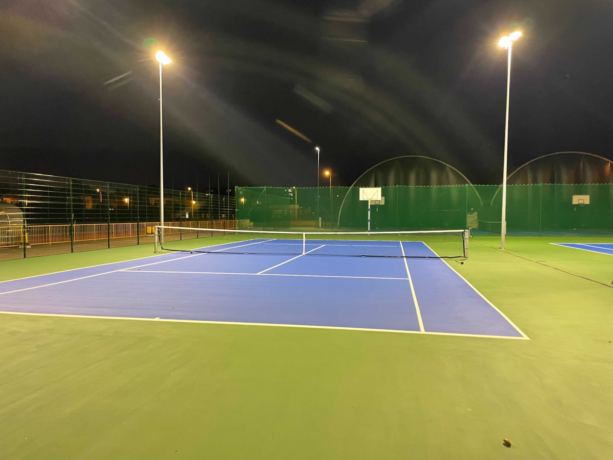 Oświetlony nocą kort tenisowy z niebiesko-zieloną nawierzchnią i białymi liniami, widoczne wysokie lampy oświetleniowe i siatka.