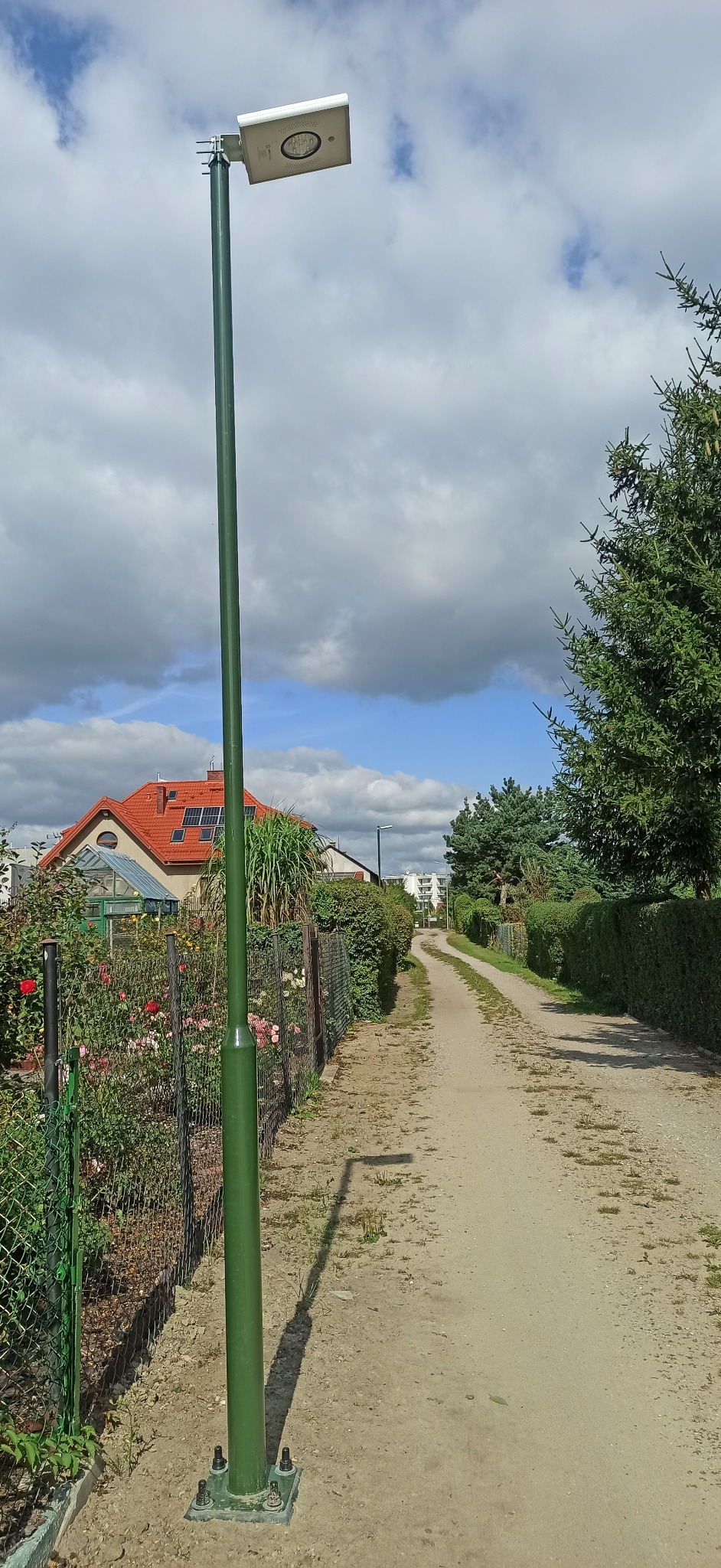 Nowoczesna, zielona lampa solarna LED oświetlająca ścieżkę w ogrodzie działkowym, widoczna z bliska na tle błękitnego nieba z chmurami i domków z czerwonymi dachami.