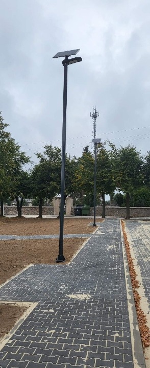 Dwie lampy solarne oświetlające park z kostki brukowej w pochmurny dzień. W tle drzewa i maszt telekomunikacyjny.