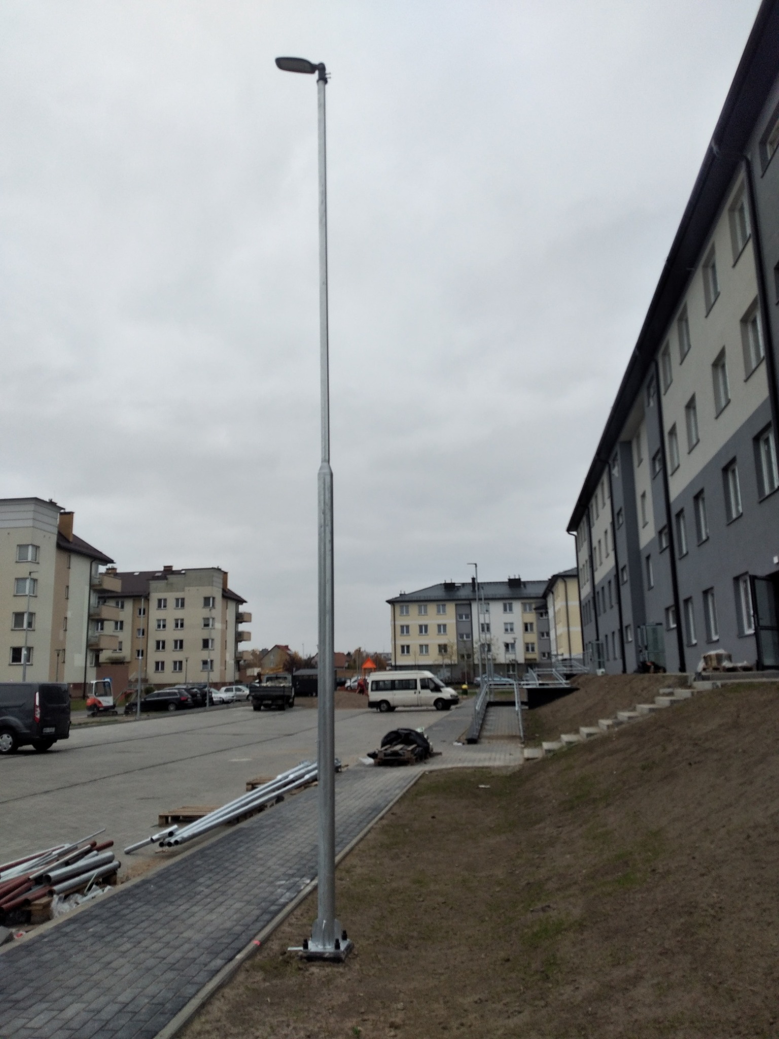 Nowo zainstalowana, wysoka, metalowa latarnia uliczna na tle bloków mieszkalnych i pochmurnego nieba. Na pierwszym planie widoczne stosy rur i kostka brukowa.