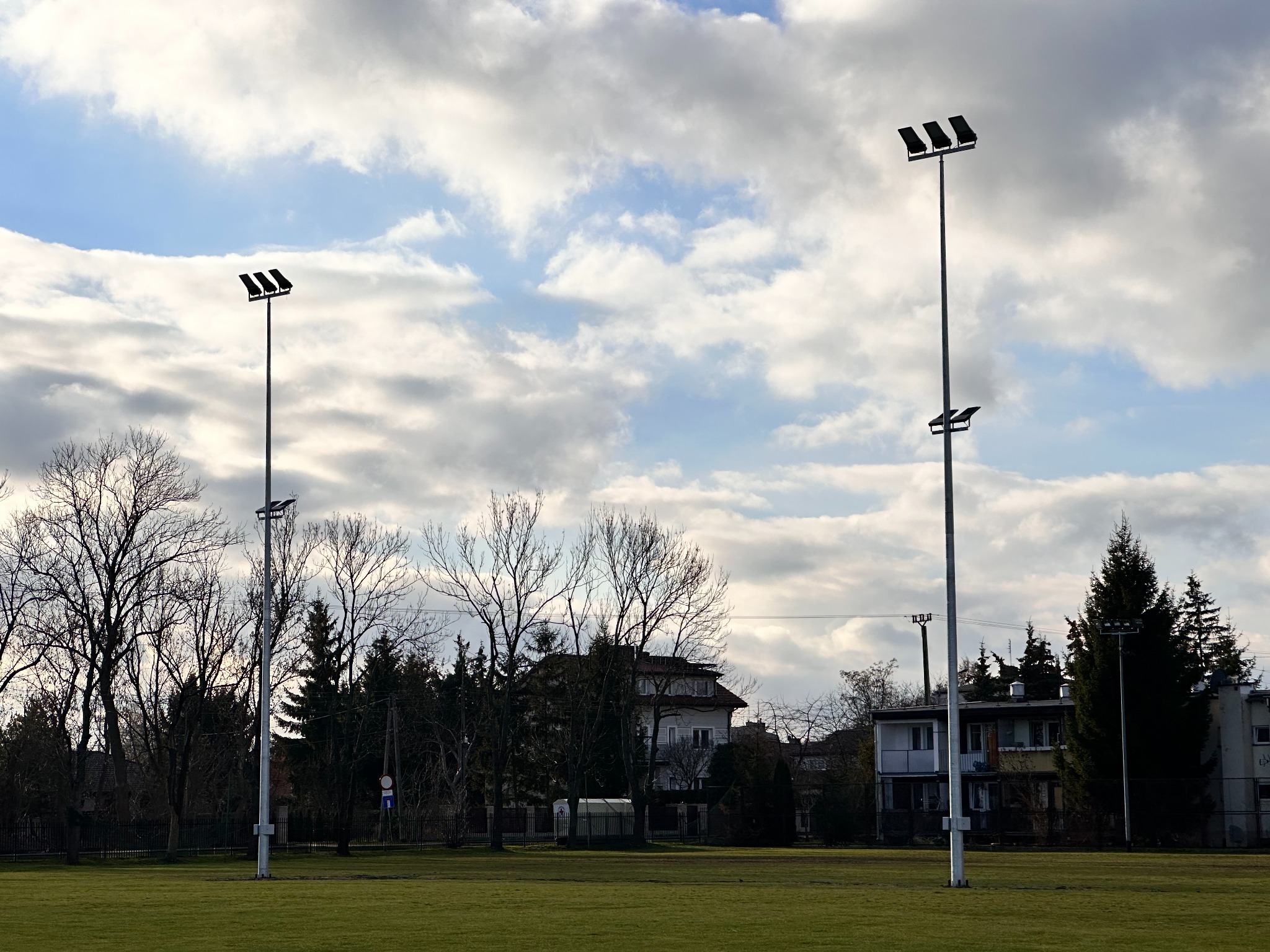 Dwa wysokie maszty oświetleniowe z trzema reflektorami każdy, ustawione na skraju zielonego boiska, z budynkami i drzewami w tle pod pochmurnym niebem.