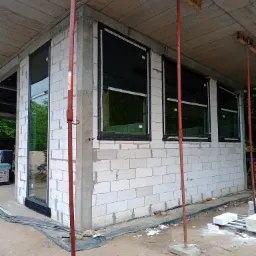 Surowy budynek z betonu komórkowego w trakcie budowy, z zamontowanymi oknami z czarnymi ramami, podparty czerwonymi stemplami budowlanymi.