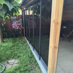 WFmontaż - Szklana zabudowa tarasu z drewnianą konstrukcją nośną, widok z zewnątrz na ogród z kwitnącymi kwiatami, widoczne przewody elektryczne na trawniku.