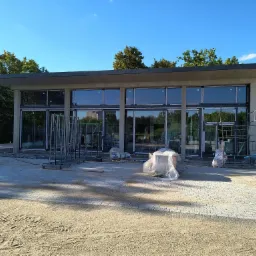 Budynek z przeszkloną fasadą w Warszawie w trakcie montażu stolarki okiennej, widoczne rusztowania, materiały budowlane i świeżo ułożona kostka brukowa.