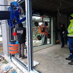 WFmontaż - Montaż dużego okna z użyciem specjalistycznego podnośnika próżniowego; widoczni pracownicy w odblaskowych ubraniach i fragment budynku w stanie surowym.