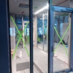 WFmontaż - Montaż aluminiowych okien z szybami zabezpieczonymi zieloną taśmą, widoczne narzędzia i elementy konstrukcyjne w surowym wnętrzu.