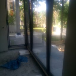 WFmontaż - Częściowo otwarte, panoramiczne okno z czarną ramą, widok na zewnątrz na drzewa i budynek, wewnątrz widoczny nieporządek budowlany, w tym niebieska plandeka i płyty.