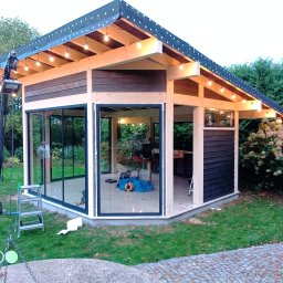 WFmontaż - Nowoczesna altana ogrodowa z przeszklonymi ścianami i drewnianą konstrukcją, oświetlona girlandą żarówek, widoczna drabina i narzędzia sugerują trwające prace wykończeniowe.