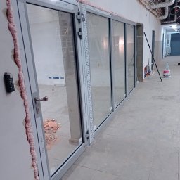 WFmontaż - Szklane drzwi przesuwne z szarymi ramami, w trakcie montażu, widoczne pianka montażowa, gruz i drabina w tle.