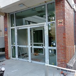 WFmontaż - Montaż konstrukcji okiennej z białych profili w budynku z cegły, widoczna drabina i narzędzia, folia ochronna na profilach.