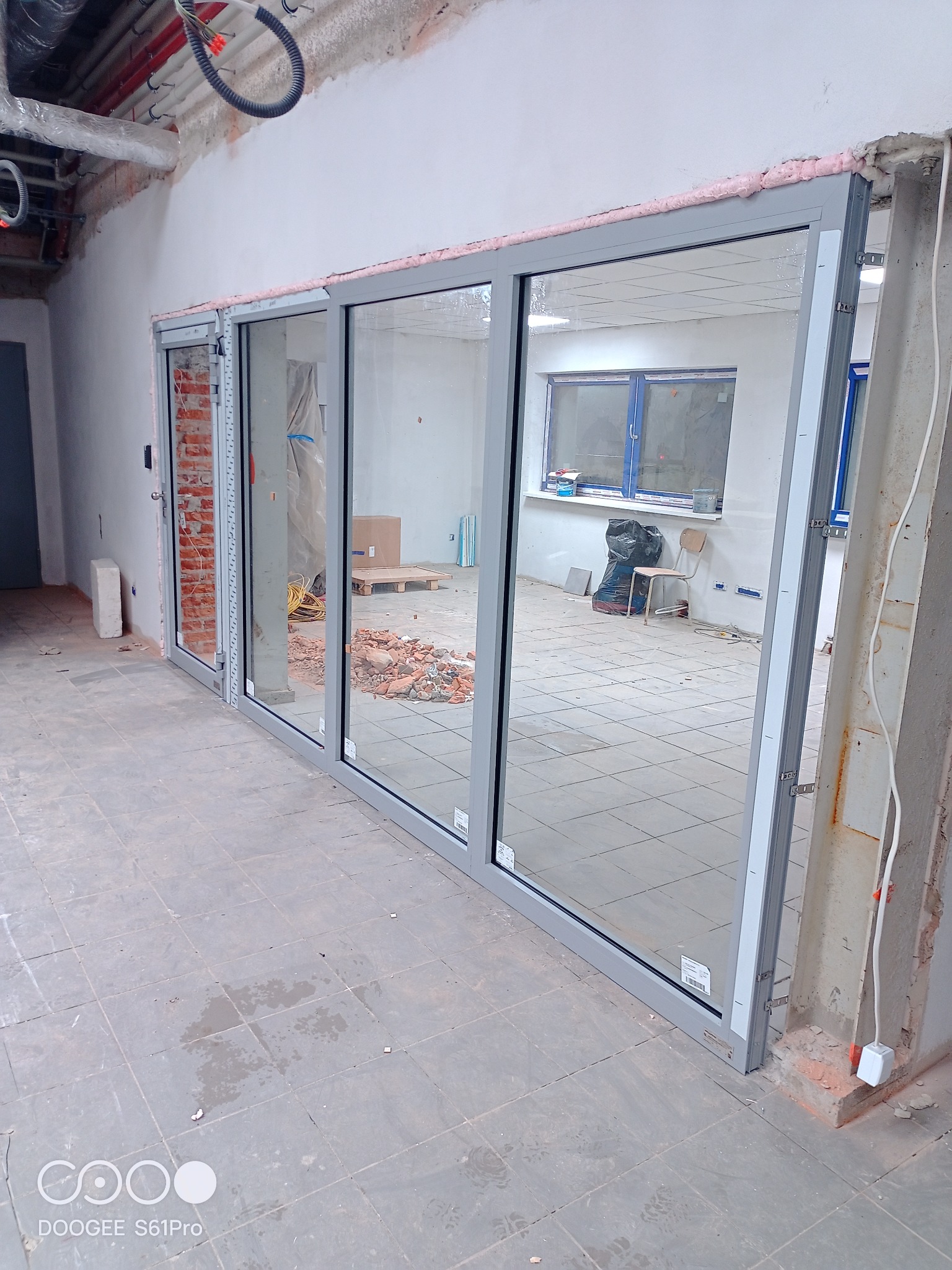 Montaż szklanej ściany działowej z aluminiową ramą w surowym wnętrzu, widoczne ślady prac budowlanych i niezabezpieczone przewody.