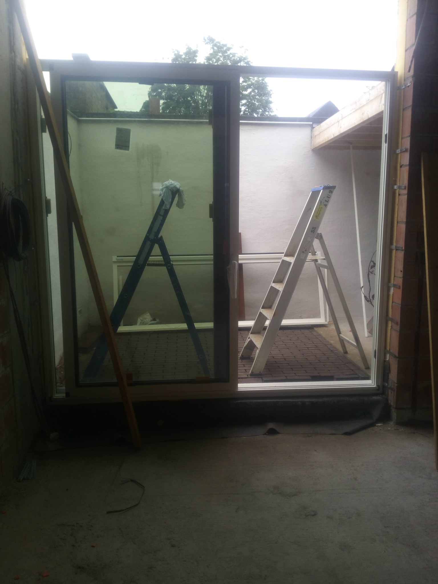 Wnętrze w trakcie montażu dużego okna przesuwnego z widokiem na patio; widoczne drabiny, ceglane ściany i elementy konstrukcyjne.