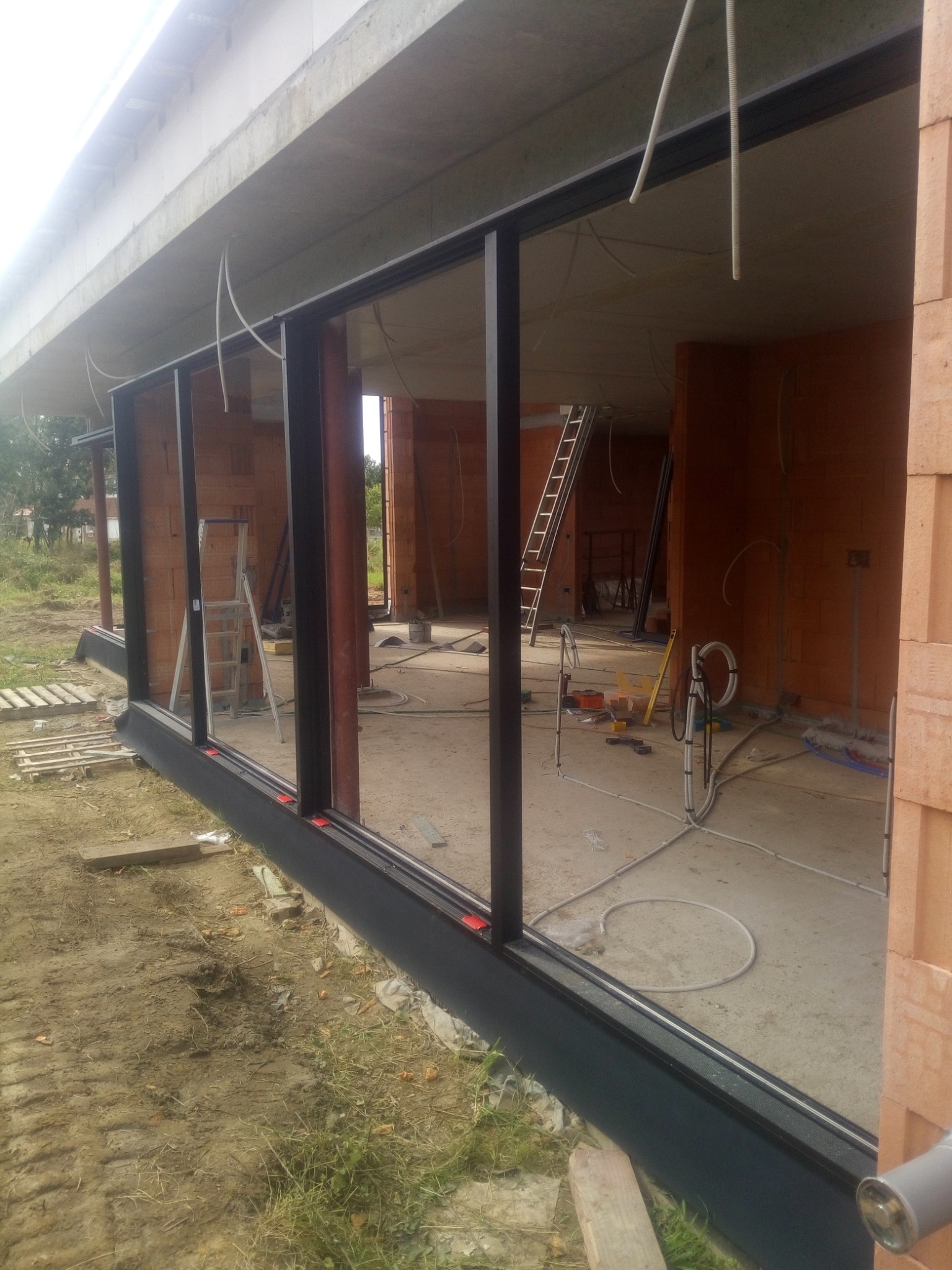 Częściowo zamontowane, panoramiczne okno przesuwne w surowym stanie, widoczne narzędzia i kable wewnątrz, ceglane ściany w tle.