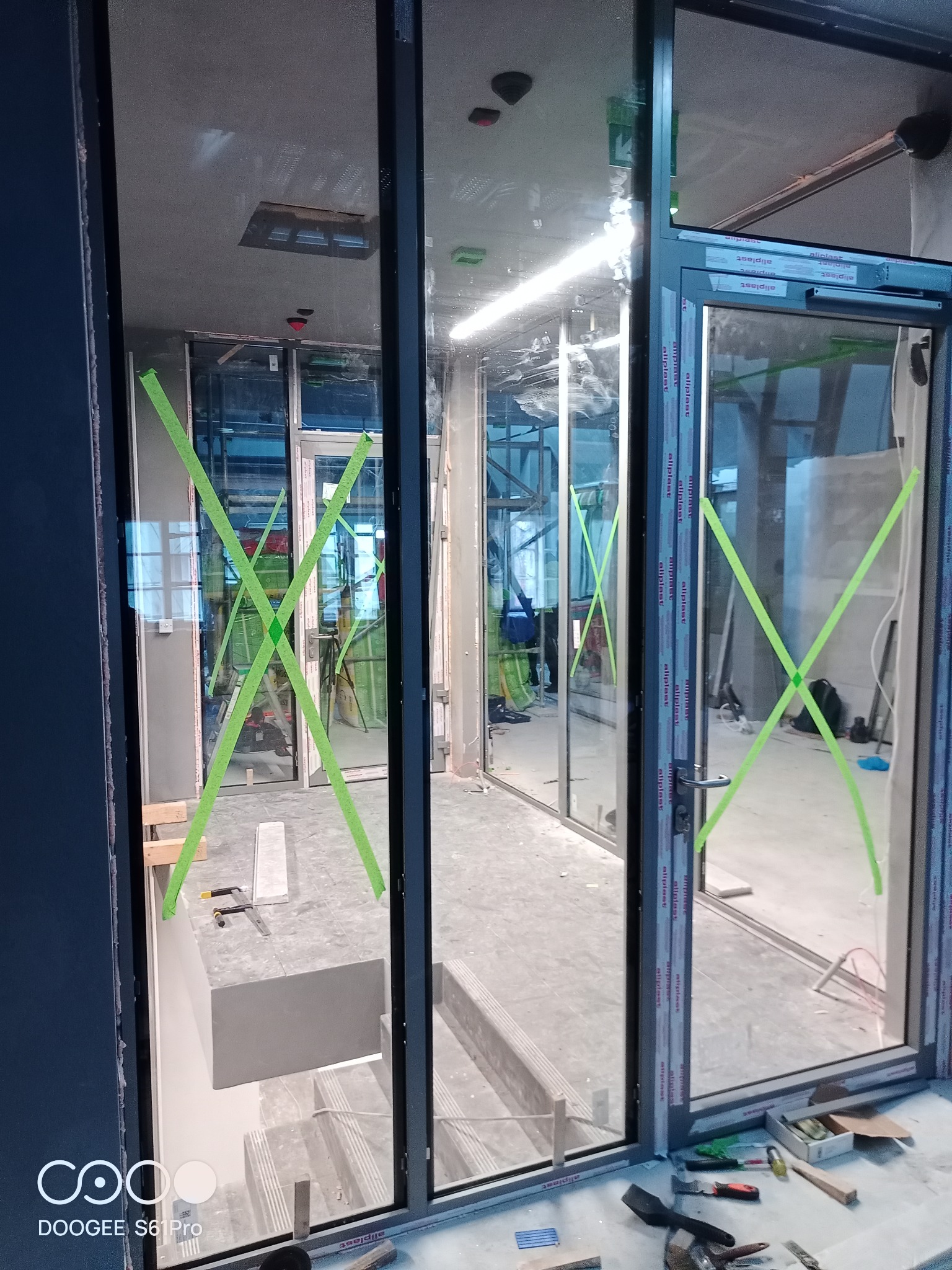 Montaż aluminiowych okien z szybami zabezpieczonymi zieloną taśmą, widoczne narzędzia i elementy konstrukcyjne w surowym wnętrzu.