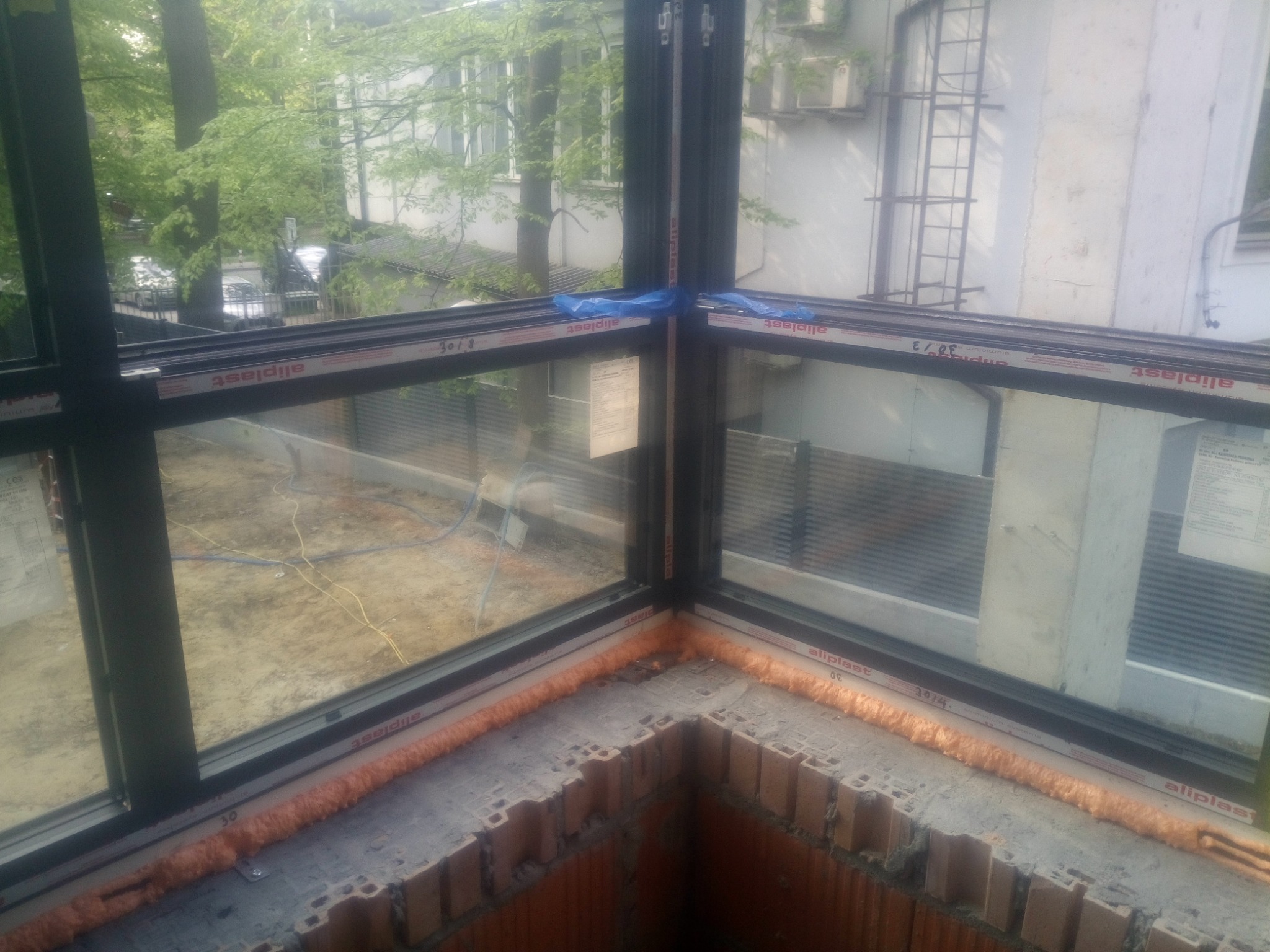 Narożne okno z czarną ramą, uszczelnione pianką montażową, widok z zewnątrz na budynek i zieleń.