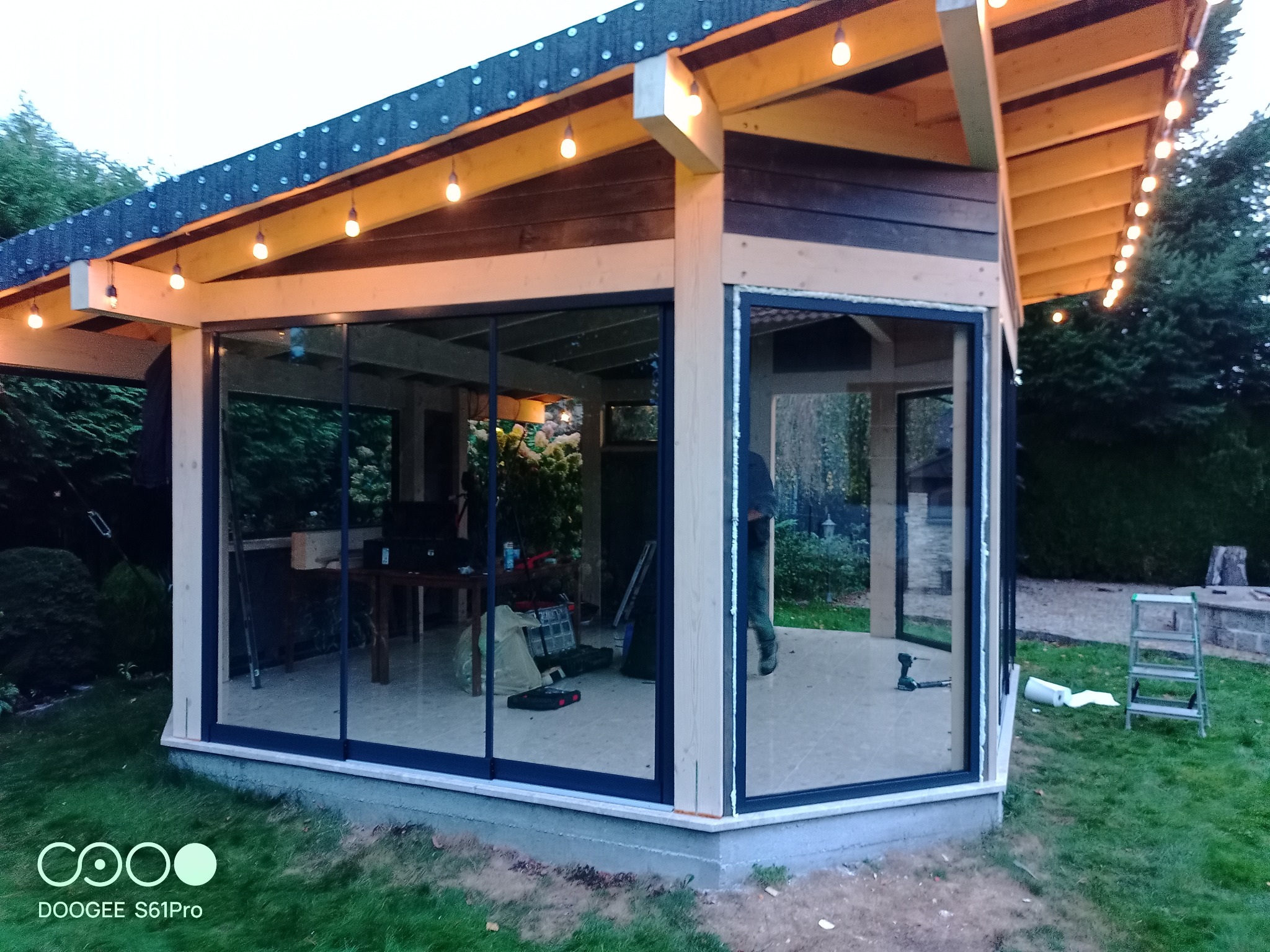 Pawilon ogrodowy z przeszklonymi ścianami w trakcie montażu, widoczne narzędzia i drabina na zewnątrz, oświetlenie sznurkowe pod dachem.