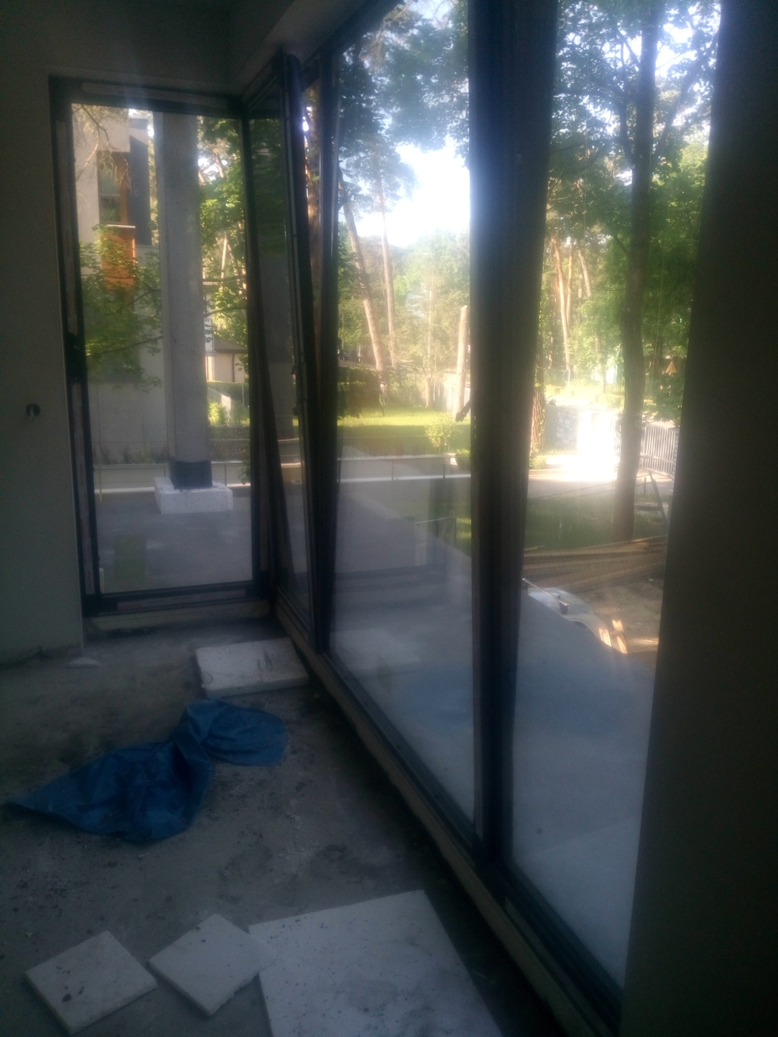 Częściowo otwarte, panoramiczne okno z czarną ramą, widok na zewnątrz na drzewa i budynek, wewnątrz widoczny nieporządek budowlany, w tym niebieska plandeka i płyty.