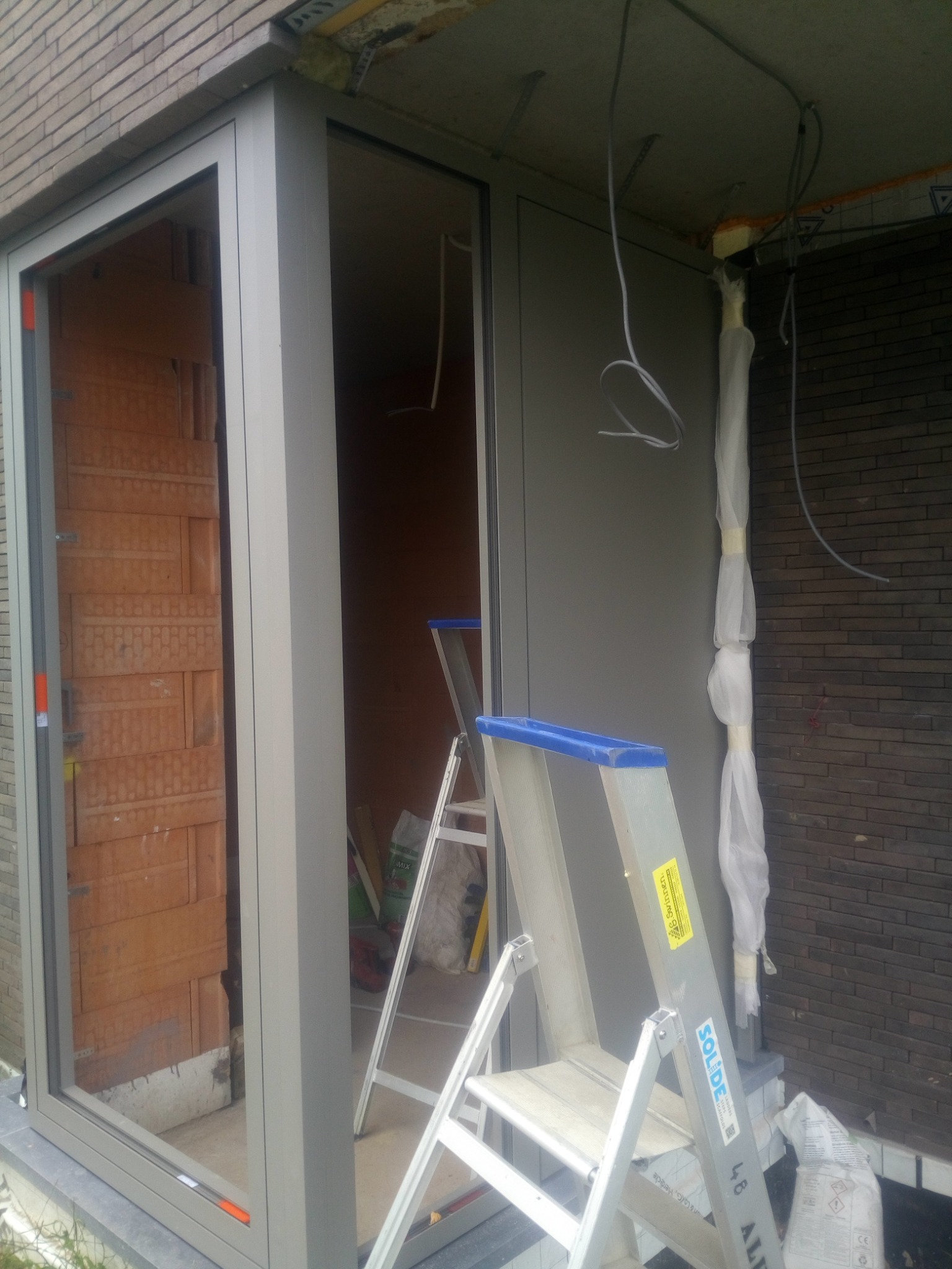 Szara rama okienna w trakcie montażu, widoczna ceglana ściana wewnątrz, drabina oparta o ścianę, elementy instalacji elektrycznej zwisające z sufitu.