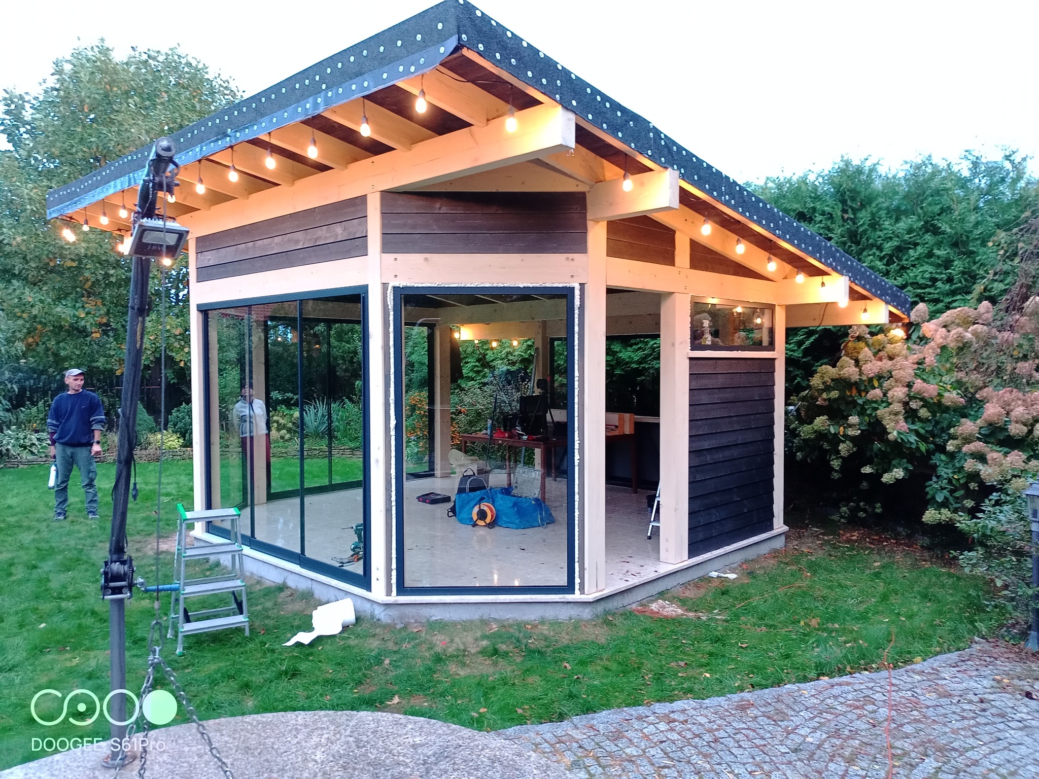 Nowoczesna altana ogrodowa z przeszklonymi ścianami i drewnianą konstrukcją, oświetlona girlandą żarówek, widoczna drabina i narzędzia sugerują trwające prace wykończeniowe.