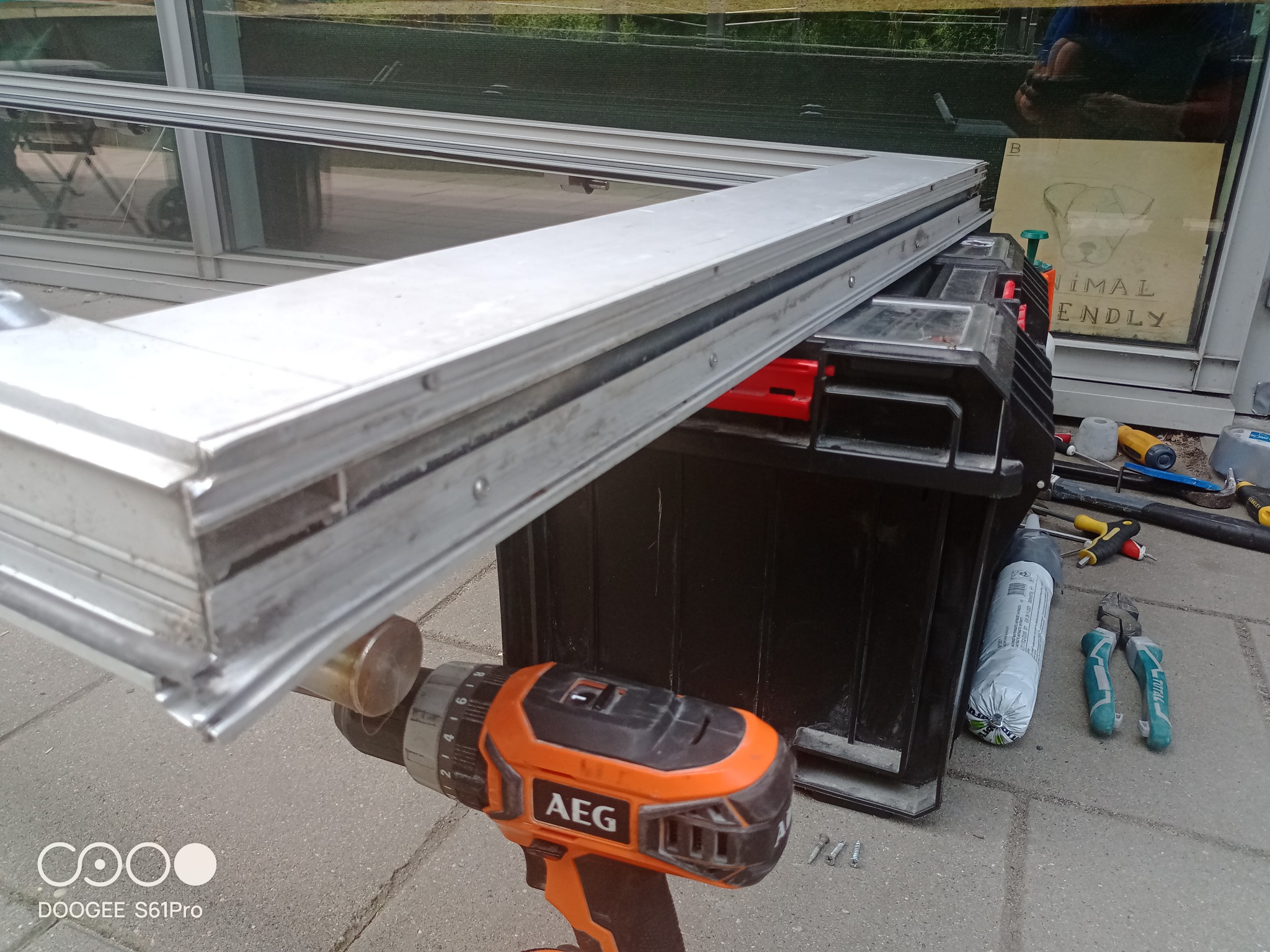 Profil aluminiowy okna na skrzynce narzędziowej, wiertarko-wkrętarka AEG obok, w tle szyba z naklejką 'Animal Friendly'.