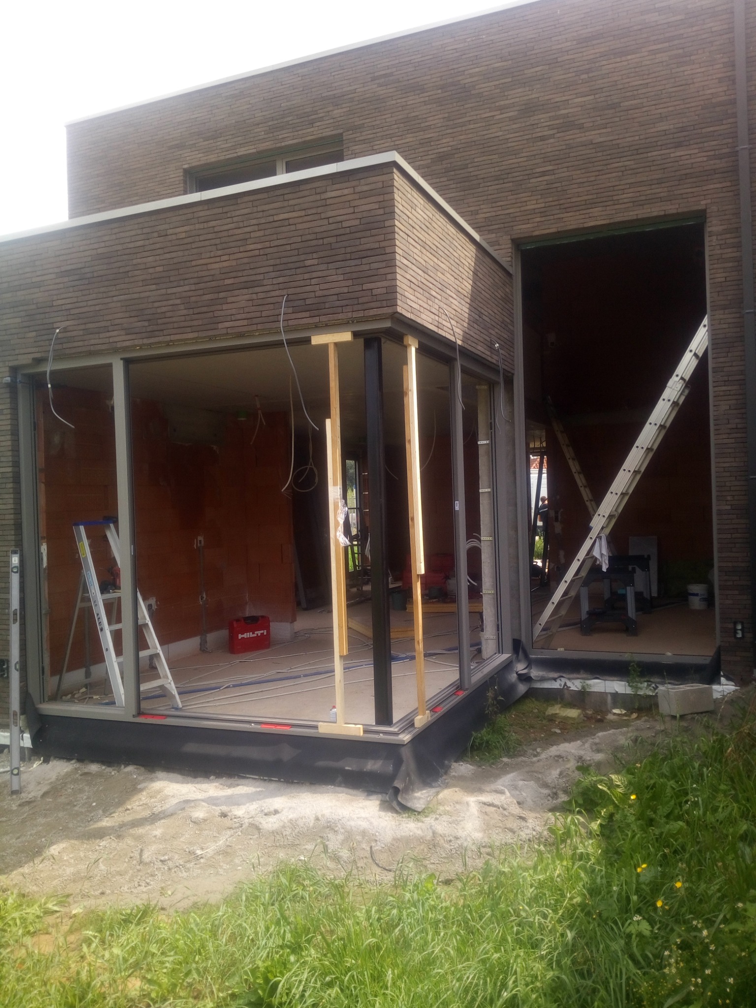 Montaż okien aluminiowych w surowym stanie, widoczne profile, drabina, narzędzia i ceglany budynek w tle.