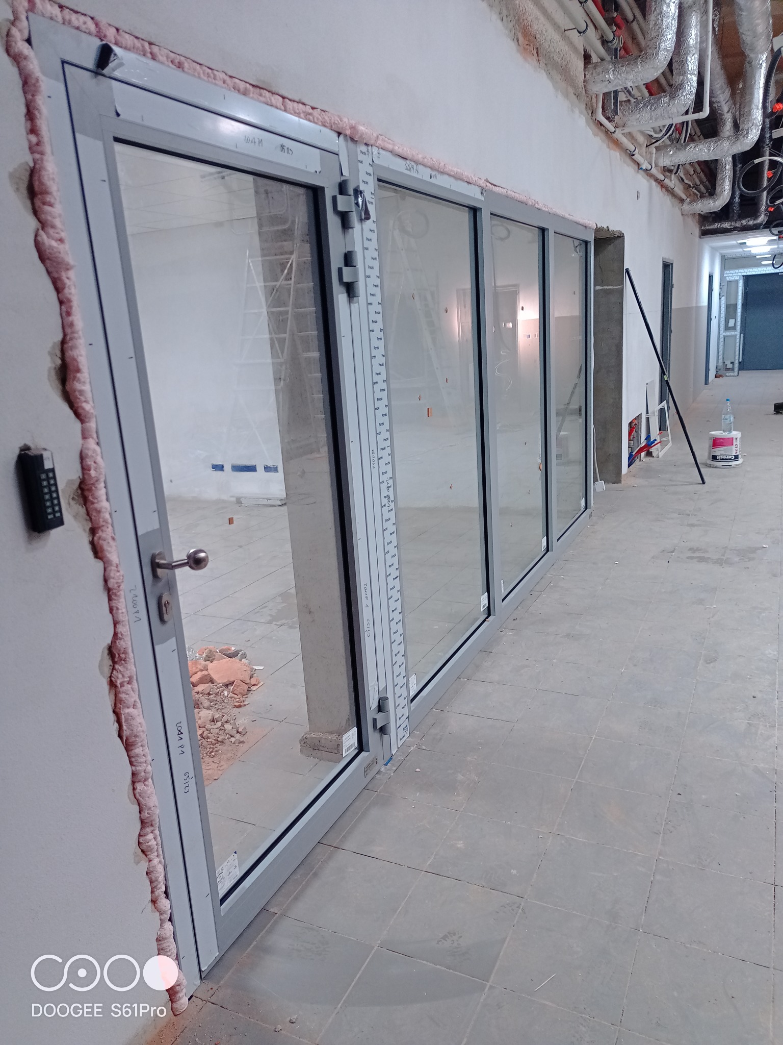 Szklane drzwi przesuwne z szarymi ramami, w trakcie montażu, widoczne pianka montażowa, gruz i drabina w tle.