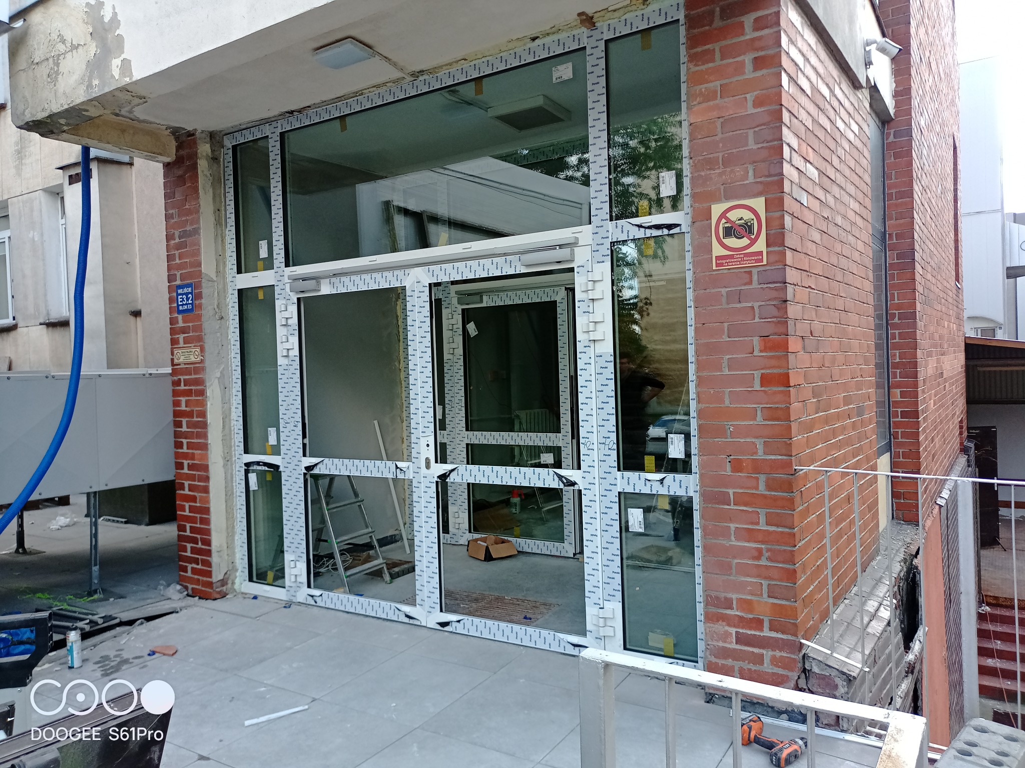 Montaż konstrukcji okiennej z białych profili w budynku z cegły, widoczna drabina i narzędzia, folia ochronna na profilach.