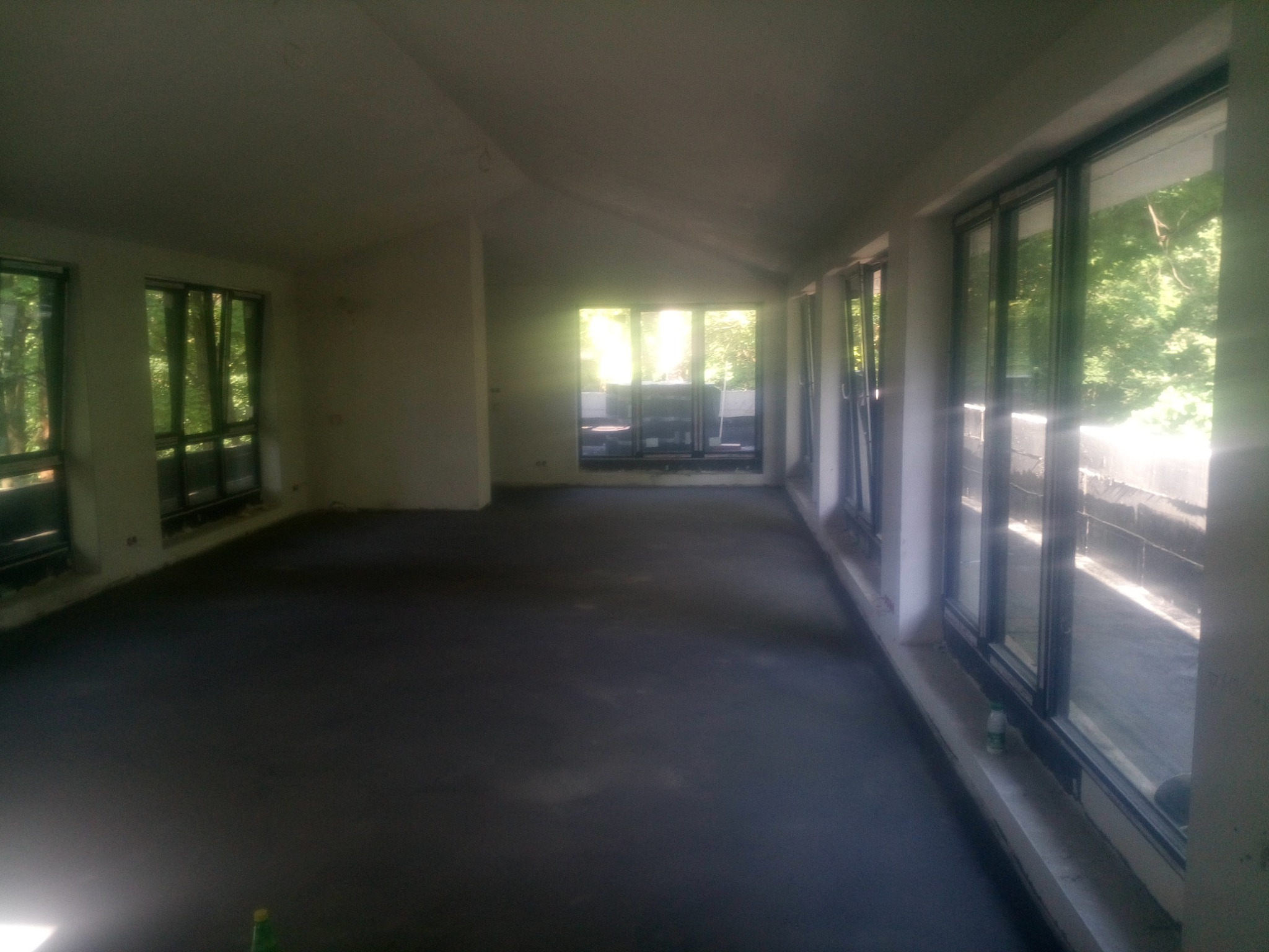 Wnętrze pomieszczenia z dużymi oknami, widok na zieleń za oknem, surowy stan wykończenia, posadzka przygotowana pod wylewkę, butelka po napoju przy oknie.