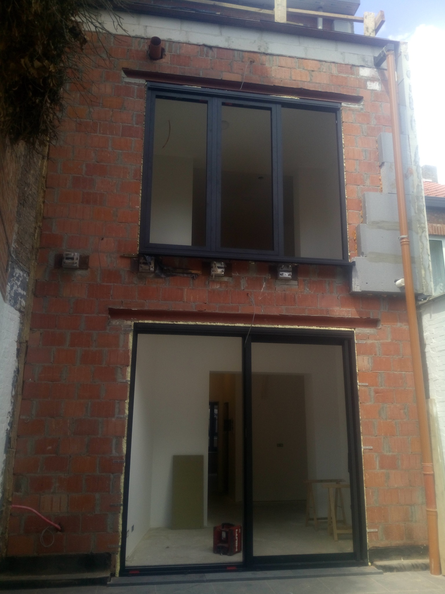 Niedokończona elewacja budynku z dwoma otworami okiennymi/drzwiowymi, widoczny mur z czerwonej cegły, stalowe belki nośne, instalacja odpływowa i narzędzia wewnątrz budynku.