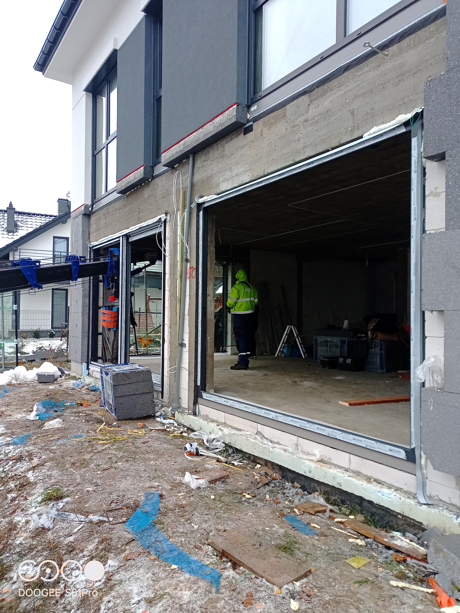 Prace przygotowawcze do osadzenia dużych okien przesuwnych w nowym budynku w Warszawie, widoczny pracownik w odblaskowej kurtce, dźwig pomocniczy i materiały budowlane.